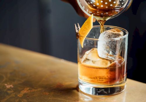 A cocktail being poured into a glass with a large ice cube and garnished with a lemon twist.