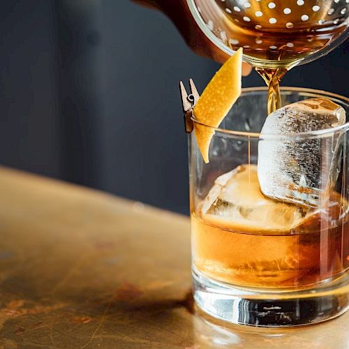 A cocktail being poured into a glass with a large ice cube and garnished with a lemon twist.