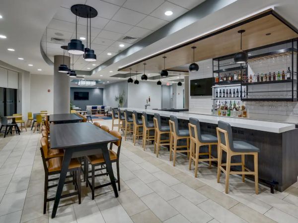 The image depicts a modern bar area with a long counter, bar stools, tables, and chairs. Bottles and glasses are displayed behind the counter.