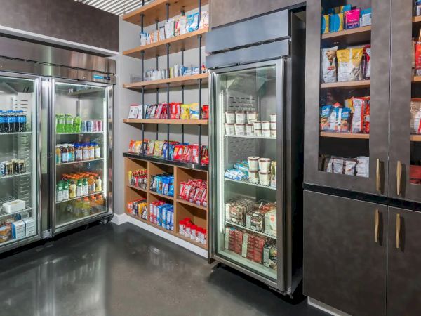The image shows a small convenience store or pantry with refrigerated units and shelves stocked with various snacks and drinks.